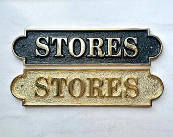 May include: Two rectangular metal plaques with rounded ends, each featuring the word STORES in raised, polished gold-tone lettering. The top plaque has a textured black background, while the bottom plaque has a textured gold-tone background.