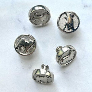 May include: Five silver-toned buttons with dog silhouettes. The buttons have a rounded shape with a raised rim and a small loop on the back for sewing. The dog designs vary, with some showing a full dog and others a profile.