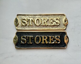 May include: Two rectangular brass store signs. One is gold with a textured surface, the other is black. Both signs have the word "STORES" in gold, raised lettering. Each sign has two screw holes.