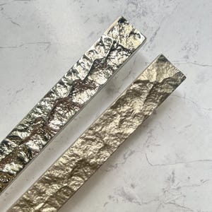 May include: Two textured metal cabinet pulls. One is silver, the other is gold. Both are rectangular with a hammered, uneven surface. The pulls are angled diagonally across the frame.