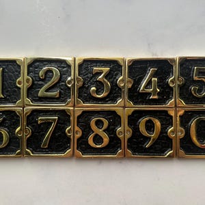 Peut inclure: Ensemble de dix numéros de maison en laiton, de 0 à 9, avec une finition dorée et un fond noir. Chaque numéro est carré avec des coins arrondis et des trous de vis pour le montage. Idéal pour l'identification de l'adresse.