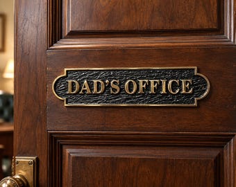 May include: A rectangular metal door sign with a black textured background and raised gold-colored lettering that reads DAD'S OFFICE. The sign features a decorative border and is mounted on a dark brown wooden door with a brass door handle.