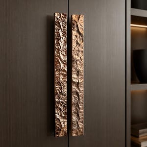 May include: Two vertical rectangular cabinet handles with a textured, rugged, bronze-colored metallic finish. The handles are mounted side-by-side on dark brown, smooth-surfaced cabinet doors, featuring a distinct uneven surface pattern.