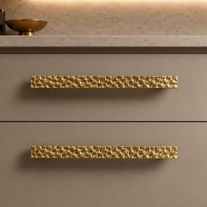 Könnte beinhalten: Zwei horizontale Schubladengriffe mit glänzend goldener Oberfläche und einer texturierten, gehämmerten Struktur. Die rechteckigen Griffe sind an einer flachen, matten, taupefarbenen Schrankfront montiert und bilden einen deutlichen Kontrast.