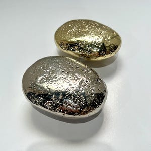 May include: Two decorative, pebble-shaped objects. One is gold-colored with a textured surface, and the other is silver-colored with a similar texture. The objects are smooth and rounded, with a metallic sheen, and are set against a white background.