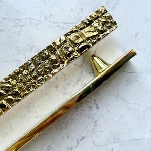 May include: Two gold-colored decorative cabinet pulls. One pull has a textured, rectangular design, while the other is a simple, straight bar with a conical end. The pulls are set against a white and gray marble background.