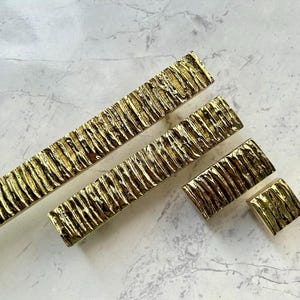 May include: A collection of gold-toned cabinet handles and knobs. The set includes long rectangular pulls and square knobs, all with a textured, ridged design. This hardware is suitable for kitchen or bathroom cabinets, adding a touch of luxury.