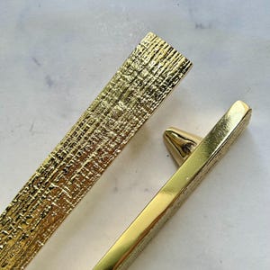 May include: Close-up of two gold-colored cabinet pulls. One pull has a textured, cross-hatched pattern, while the other is smooth and rectangular. The pulls are likely for furniture or cabinetry.
