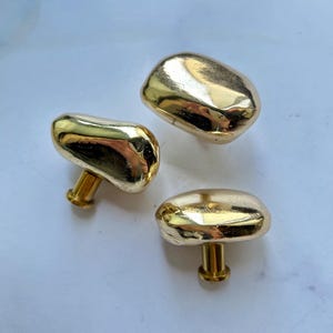 May include: Three gold-colored cabinet knobs with a smooth, irregular, pebble-like shape. Each knob features a polished, reflective metallic surface and a cylindrical base for attachment, showcasing a rounded, organic form without sharp edges.