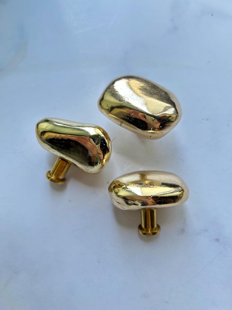 May include: Three gold-colored cabinet knobs with a smooth, irregular, pebble-like shape. Each knob features a polished, reflective metallic surface and a cylindrical base for attachment, showcasing a rounded, organic form without sharp edges.