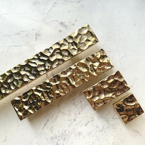 May include: A collection of gold-colored cabinet pulls and knobs with a hammered texture. The hardware is arranged on a white and gray marble surface. The pulls vary in size, with the longest measuring approximately 12 inches.