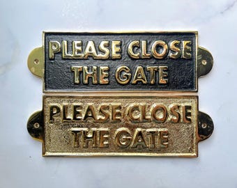 Op de afbeelding: Twee rechthoekige metalen borden met de tekst "PLEASE CLOSE THE GATE" in gouden letters. Eén bord is zwart, de andere is goudkleurig. Beide hebben afgeronde hoeken en montagegaten.