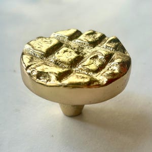 以下が含まれることがあります： 丸い上面にテクスチャ加工された幾何学模様が施された、金色のキャビネットノブ。ノブには、取り付け用の短い円筒形のベースが付いています。仕上げは光沢があり反射性があり、真鍮または同様の金属を示唆しています。