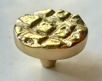 May include: A gold-colored cabinet knob with a textured, geometric design on the round top. The knob has a short, cylindrical base for mounting. The finish is shiny and reflective, suggesting a brass or similar metal.