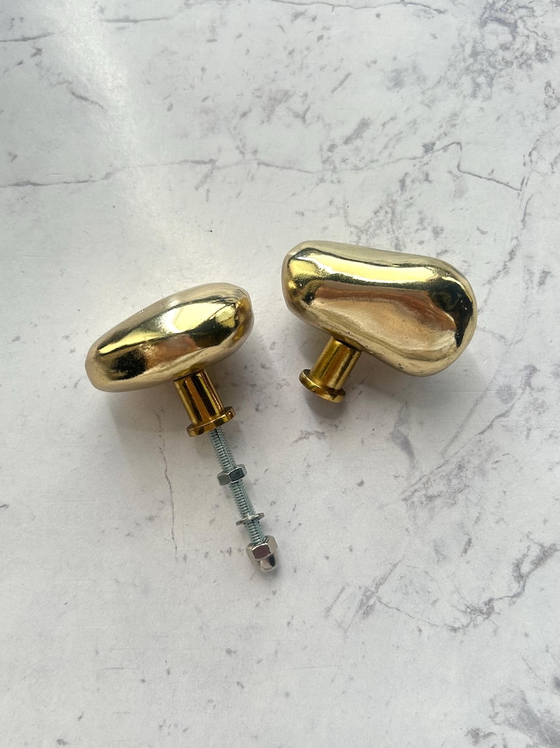 May include: Two gold-colored cabinet knobs with a smooth, organic, pebble-like shape. One knob features a long threaded metal bolt extending from the base, equipped with two small metal nuts and a washer for secure installation on furniture.