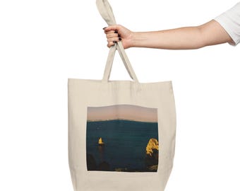 Puede incluir: Un bolso tote beige con una imagen rectangular de un paisaje marino. La imagen muestra un océano azul oscuro con una formación rocosa y una costa distante. El bolso es sostenido por la mano de una persona.