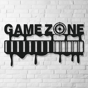 Może przedstawiać: Czarna metalowa grafika ścienna z napisem "GAME ZONE" i symbolem tarczy. Projekt zawiera stylizowany kształt kontrolera do gier z efektem spływającej farby, zamontowany na białej ceglanej ścianie.