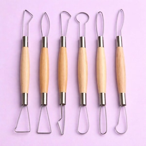 Puede incluir: Un juego de seis herramientas de cerámica con mangos de madera y lazos y cuchillas de acero inoxidable. Las herramientas están dispuestas en una fila sobre un fondo morado claro. Cada herramienta tiene un extremo de forma diferente para esculpir y dar forma a la arcilla.