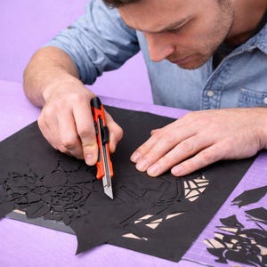 May include: A person using a red and black utility knife to cut intricate designs from a sheet of black paper. The paper features floral and geometric patterns. The person is wearing a blue denim shirt.