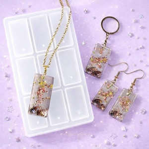 May include: A jewellery set comprising a pendant necklace, earrings, and a keyring. Each item has a rectangular shape with a clear resin design, incorporating seashells and gold flakes. The set is displayed on a lavender surface with a white mould.