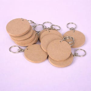 May include: A selection of blank, circular wooden keyrings with silver key rings. The keyrings are a light brown colour and are stacked and scattered across a light purple surface. The key rings are attached through a small hole at the top of each keyring.