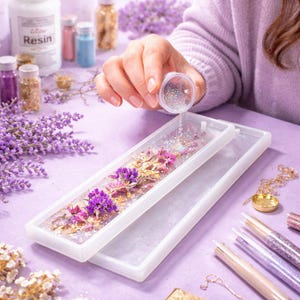 May include: A white rectangular resin mold with dried flowers and glitter. A hand is pouring glitter into the mold. Jars of resin, glitter, and dried flowers are in the background. The theme is crafting and DIY projects.