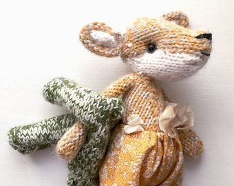 Peut inclure: Un jouet en peluche renard tricoté portant une robe à fleurs jaunes et blanches et un pull rayé vert et blanc. Le renard a des yeux bruns et un museau blanc.