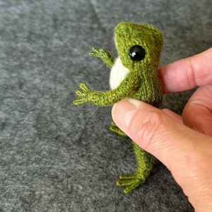 May include: A handmade green frog figurine with large black eyes and a white belly. The frog is made of a textured yarn and has outstretched arms and legs, giving it a friendly appearance. The frog is a whimsical, handcrafted item.