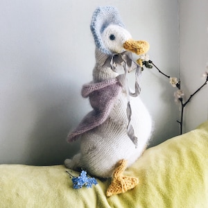 以下が含まれることがあります： 黄色の表面に青い花が咲いている上に、薄い青色の帽子と紫色のショールをつけた白い編まれたガチョウが座っています。