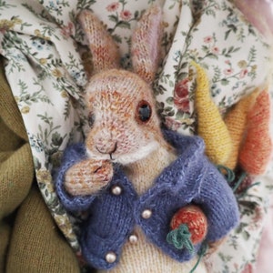 May include: A knitted brown rabbit wearing a blue cardigan with buttons and holding a carrot. The rabbit is sitting on a floral fabric background.