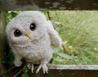 May include: A white knitted owl with black eyes and a yellow beak, perched on a wooden fence.