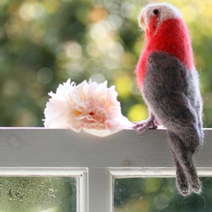 May include: A pink and grey knitted bird figurine perched on a windowsill with a pink flower in the foreground.