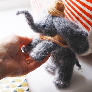 Puede incluir: Un juguete de elefante de punto gris que lleva un gorro y una bufanda de punto amarillo. El elefante tiene ojos azules y pies rosas.