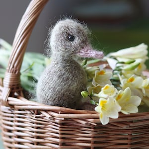Puede incluir: Un patito de punto gris se sienta en una cesta de mimbre con flores de freesia blancas y amarillas.