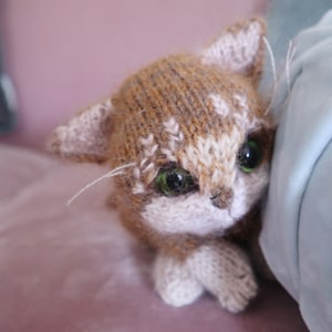 May include: A brown and white knitted cat toy with green eyes. The cat is lying on its side with its head resting on a blue surface.