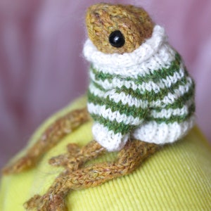 May include: A knitted frog wearing a green and white striped sweater with a white turtleneck. The frog is sitting on a yellow surface.