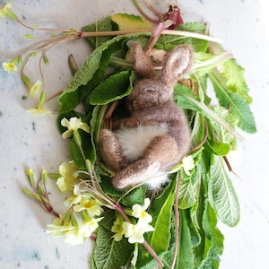 Może przedstawiać: Ręcznie wykonana figurka śpiącego królika z brązowym futrem i białym brzuchem, osadzona wśród zielonych liści i żółtych kwiatów. Królik leży, wyglądając na spokojnego. Aranżacja nawiązuje do naturalnego, wiosennego motywu.