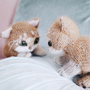 May include: Two knitted cat toys, one brown and white and one tan and white, are facing each other on a light blue surface. The cats have green eyes and white fur on their faces.