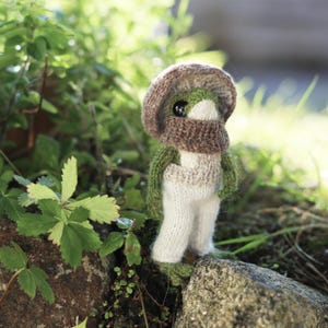 Peut inclure: Un jouet en peluche en forme de champignon crochet&eacute; avec un chapeau brun et un corps vert et blanc. Le jouet est debout sur un rocher avec de la v&eacute;g&eacute;tation verte en arri&egrave;re-plan.