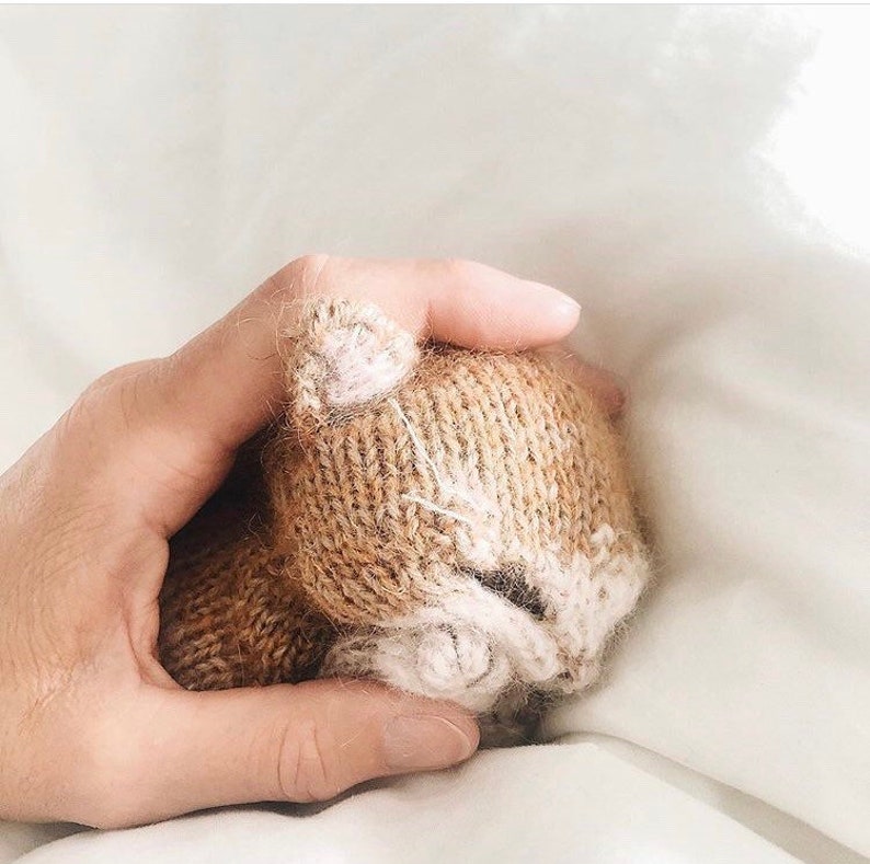 May include: A hand holding a small knitted cat toy. The cat is brown with white accents and has a sleepy expression.