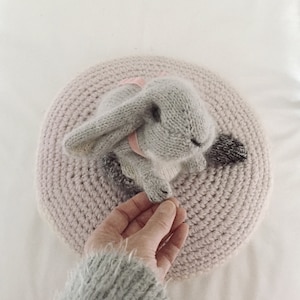 May include: A gray knitted bunny rabbit toy with a pink ribbon around its neck, sitting on a pink crocheted round cushion.