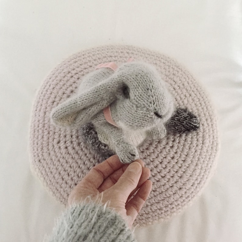 May include: A gray knitted bunny rabbit toy with a pink ribbon around its neck, sitting on a pink crocheted round cushion.
