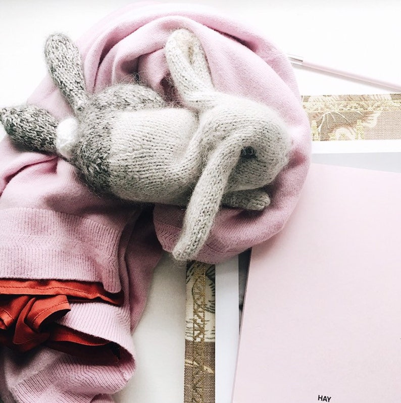 May include: A gray knitted bunny rabbit toy with long ears, lying on a pink knitted scarf. The scarf is folded and draped over a pink book with the word "HAY" printed on the cover.