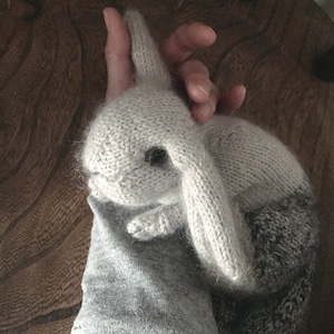 May include: A gray and white knitted bunny rabbit toy with a fluffy texture. The bunny is lying on a gray sleeve.