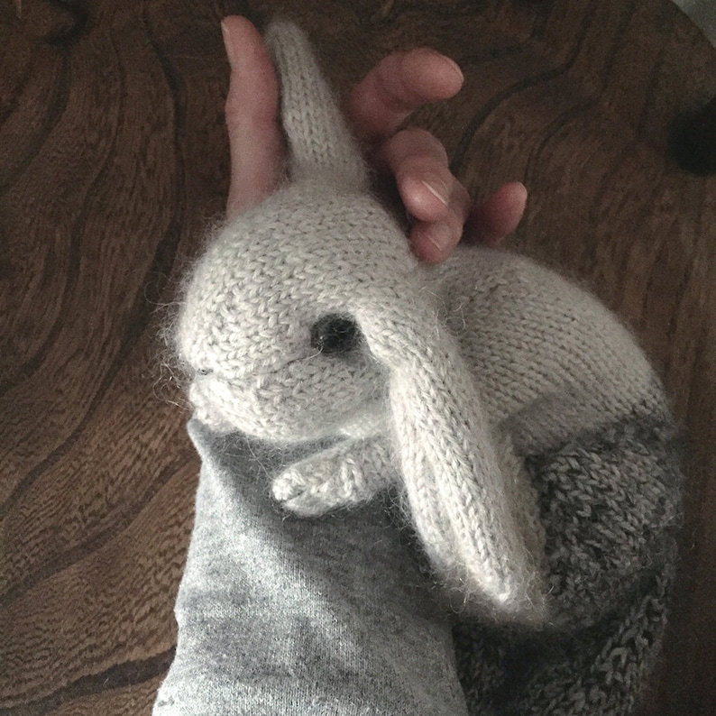 May include: A gray and white knitted bunny rabbit toy with a fluffy texture. The bunny is lying on a gray sleeve.