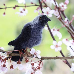 May include: A black and gray needle felted bird perched on a branch with white blossoms. The bird has a small, round body and a long, thin beak.