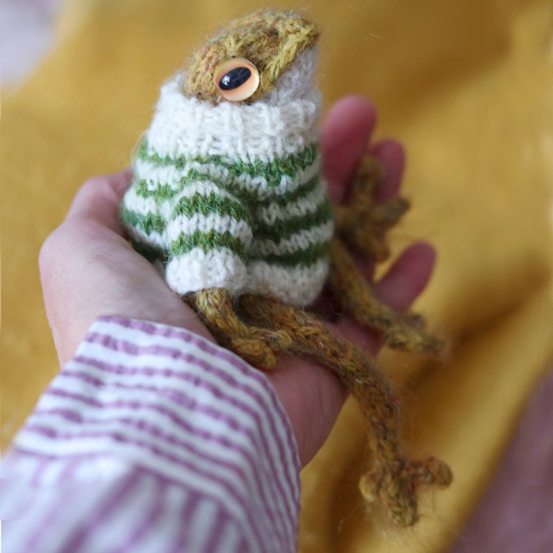 May include: A knitted frog wearing a green and white striped sweater. The frog has brown yarn legs and a yellow body.