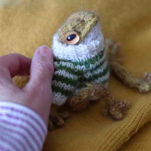 May include: A knitted frog wearing a green and white striped sweater with a white collar. The frog has a brown body and a yellow head with a single large eye.