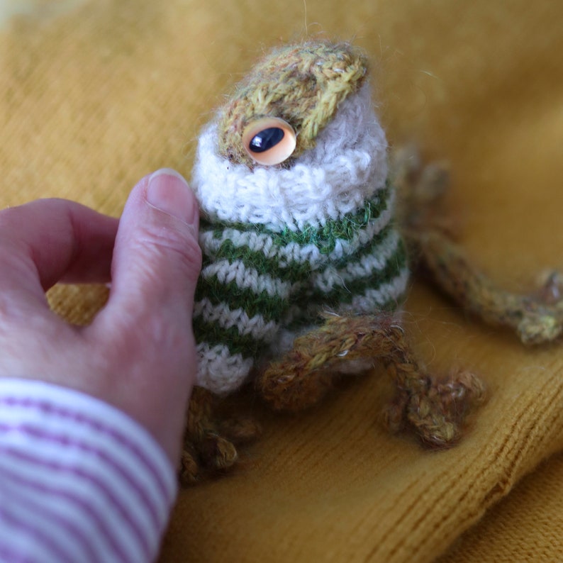 May include: A knitted frog wearing a green and white striped sweater with a white collar. The frog has a brown body and a yellow head with a single large eye.