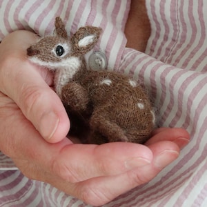 以下が含まれることがあります： 手作りの茶色と白のニット鹿の置物。白い斑点があり、すぼまった手で持たれています。背景はピンクと白のストライプの生地です。